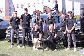 Gruppenfoto des Teams vom Autohaus Peter beim Reitturnier in Immenrode: Acht Teammitglieder in schwarzen Polohemden posieren vor ausgestellten Mercedes-Benz Fahrzeugen, im Hintergrund reiten Teilnehmer auf braunen Pferden über den Turnierplatz. (Foto: Autohaus Peter)