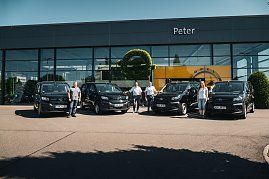 Vor einem modernen Autohaus mit dem Namen „Peter“ stehen vier schwarze Opel-Transporter in einer Reihe. Vier Personen  drei Männer und eine Frau  posieren jeweils neben den Fahrzeugen. An der Glasfassade ist das Opel-Logo und ein gelber Hintergrund zu sehen. (Foto: Autohaus Peter Gruppe)