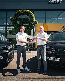 Vor dem Autohaus „Peter“ stehen zwei Männer zwischen zwei schwarzen Opel-Fahrzeugen. Einer der Männer überreicht dem anderen einen Autoschlüssel, beide lächeln und geben sich die Hand. Im Hintergrund ist das Opel-Logo aus grünem Pflanzenmaterial an der Glasfassade zu sehen. Die Szene wirkt freundlich und sonnig. (Foto: Autohaus Peter Gruppe)