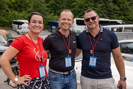 Christina Dost, Carsten Dost und Enrico Dost stehen Arm in Arm vor geparkten Fahrzeugen, tragen Namensschilder und rote Lanyards. Im Hintergrund sind Busse, Autos und grüne Bäume zu sehen  die Szene bei der Rallye wirkt fröhlich. (Foto: AUTOHAUS)