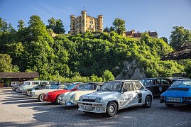 Reihe von klassischen Autos, darunter ein weißer Renault 5 Turbo mit Startnummer 52, ein roter und ein cremefarbener Porsche, die auf einem Parkplatz vor einer grünen bewaldeten Anhöhe stehen. Im Hintergrund thront das gelbe Schloss Hohenschwangau in Bayern unter blauem Himmel. Die Szene wirkt sonnig und freundlich. (Foto: AUTOHAUS)
