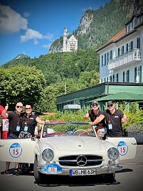 Das Team vom Autohaus Peter steht vor einem weißen Mercedes-Benz Oldtimer mit Startnummer 15. Zwei Personen stehen links neben dem Auto, zwei rechts. Alle tragen schwarze Shirts, Sonnenbrillen und zeigen Daumen nach oben. Im Hintergrund ist Schloss Neuschwanstein auf einem bewaldeten Berg zu sehen, daneben ein weißes Gebäude mit grünen Fensterläden. Sonniger, klarer Tag. (Foto: AUTOHAUS)