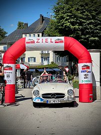 Weißer Mercedes-Benz Oldtimer mit Startnummer 15 fährt unter einem roten Zielbogen mit der Aufschrift ZIEL bei einer Rallye ein. Im Auto jubeln die Insassen mit erhobenen Armen. Menschen stehen rechts und links, klatschen und feiern. Im Hintergrund sind Gebäude mit Balkonen und Bäume zu sehen. Sonniger Tag, festliche Stimmung. (Foto: AUTOHAUS)
