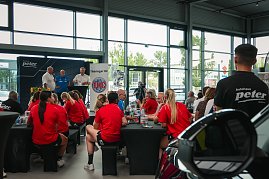 Helmut Peter, Trainer Herbert Müller und Robert Böhm (von links nach rechts) stehen auf einer Bühne beim Medien-Tag des Thüringer HC in Erfurt. Im Vordergrund sitzen Spielerinnen in roten Trikots an Tischen und hören zu. Hinter den Männern steht ein THC-Banner und durch die großen Glasfenster sind Autos und Bäume zu sehen. (Foto: Autohaus Peter)
