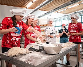 Spielerinnen des Thüringer HC in roten und weißen Trikots mit weißen Bäckermützen stehen in der Backstube der Bäckerei Helbing. Sie kneten und formen Teig, lachen und haben sichtlich Spaß. Im Vordergrund liegt Teig auf dem Tisch, daneben eine Schüssel mit Mehl. Eine Frau im Hintergrund filmt die Szene. (Foto: Autohaus Peter)