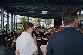 Blick von hinten auf Lilly Hecker und Geschäftsführer Andreas Peter, während sie vor den neuen Auszubildenden sprechen. Viele junge Menschen sitzen in Reihen und hören zu. (Foto: Autohaus Peter)