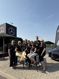 Gruppenfoto des Teams Peter beim Leichtathletikmeeting in Sondershausen. Sieben Personen tragen schwarze Polos mit Logo. Zwei sitzen auf hohen Barhockern, die anderen stehen daneben. Im Hintergrund sind ein schwarzer Mercedes-Cube mit großem Logo sowie ein schwarzer Mercedes CLA zu sehen. Alle lächeln und posieren fröhlich in der Sonne. (Foto: Autohaus Peter)