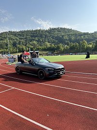 Graues Mercedes-Cabrio fährt auf die Tartanbahn ins Stadion ein. Zwei Personen sitzen oben auf der Rückbank. Im Hintergrund Zuschauertribünen und grüne Hügel. (Foto: Autohaus Peter)
