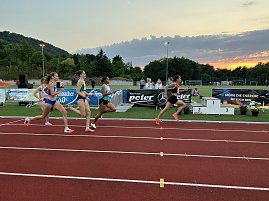 Feld von Läuferinnen sprintet nebeneinander auf der Tartanbahn. Im Hintergrund Zielbereich mit Bannern und Zuschauertribüne, die Sonne geht unter. (Foto: Autohaus Peter)