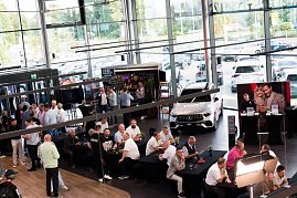 Eine große Halle mit vielen Gästen, die an schwarzen Tischen sitzen oder an Stehtischen stehen. Ein weißer Mercedes ist ausgestellt, dahinter eine Bar und ein Bildschirm mit dem Mercedes-Logo. (Foto: Buchardt | Autohaus Peter )