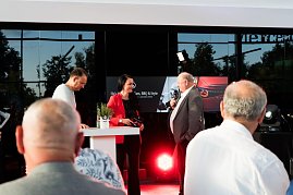 Christina Dost im roten Blazer spricht mit Helmut Peter im grauen Anzug. Beide stehen auf einer Bühne im Autohaus, links daneben, ebenfalls auf der Bühne, Andreas Peter. Das Banner „Hes Mercedes  Cars, BBQ & Style“ ist im Hintergrund zu sehen. (Foto: Buchardt | Autohaus Peter )