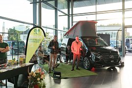 Ein schwarzer Mercedes-Camper mit geöffnetem Aufstelldach steht auf grünem Kunstrasen im Autohaus. Daneben zwei Männer, einer zeigt den Daumen hoch. Links im Bild ein Verkaufsstand mit Prospekten und Produkten. (Foto: Buchardt | Autohaus Peter )