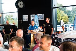 Zwei Männer stehen an einem kleinen Tisch, einer trägt ein blaues Hemd mit „DTZ“-Logo, der andere ein schwarzes Polohemd. Sie präsentieren Produkte, im Hintergrund sind ein schwarzer Mercedes und eine große Uhr zu sehen. (Foto: Buchardt | Autohaus Peter )