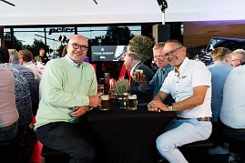 Drei Männer sitzen an einem Stehtisch mit Getränken und lachen in die Kamera. Viele weitere Gäste sitzen im Hintergrund an Tischen. (Foto: Buchardt | Autohaus Peter )