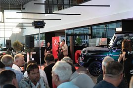 Blick in die Eventhalle mit vielen Gästen im Vordergrund. Auf der Bühne stehen Christina Dost im roten Blazer und ein Mann in beiger Jacke, daneben ein schwarzer Mercedes-Geländewagen. (Foto: Buchardt | Autohaus Peter )