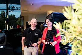 Eine Frau im roten Blazer (Christina Dost) und ein Mann im schwarzen Polohemd (Carsten Dost) stehen nebeneinander und lächeln. Beide halten Moderationskarten, im Hintergrund ein Mercedes-Geländewagen. (Foto: Buchardt | Autohaus Peter )