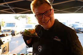 Ein Koch mit Brille und schwarzer Schürze mit Mercedes-Logo hält lächelnd ein belegtes Sandwich in die Kamera. Im Hintergrund ein Foodtruck und Fahrzeuge im Sonnenlicht. (Foto: Buchardt | Autohaus Peter )