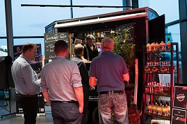 Mehrere Männer stehen an einem Gin-Stand mit schwarzem Verkaufswagen. Auf Schildern steht „Luzifer Gin“ und „Gin-Cocktails“. Regale mit Flaschen sind daneben aufgestellt. (Foto: Buchardt | Autohaus Peter )