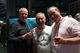 Drei Männer stehen nebeneinander und lachen in die Kamera. Einer trägt ein weißes Mercedes-Benz-T-Shirt, die anderen dunkle Hemden. Alle haben Getränke in der Hand. (Foto: Buchardt | Autohaus Peter )