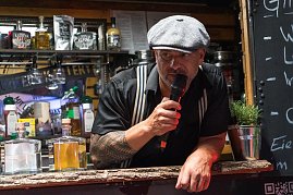 Ein Mann mit Schiebermütze und Tattoos spricht ins Mikrofon an einem Gin-Stand. Vor ihm mehrere Flaschen verschiedener Spirituosen. (Foto: Buchardt | Autohaus Peter )