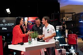 Christina Dost im roten Blazer interviewt einen jungen Mann im weißen Mercedes-AMG-Petronas-Shirt. Beide lachen. Dahinter ein Stand mit Mercedes-Merchandise. (Foto: Buchardt | Autohaus Peter )