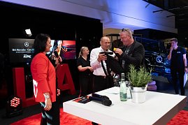 Ein Mann in weißem Hemd und ein weiterer in schwarzer Kleidung packen gemeinsam ein Geschenk aus. Links steht Christina Dost im roten Blazer mit Mikrofon. (Foto: Buchardt | Autohaus Peter )