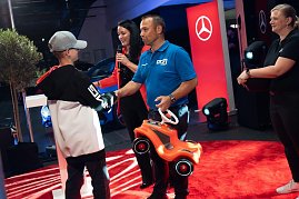 Ein Junge mit Mercedes-Cap bekommt ein rotes Bobbycar von einem Mann im blauen DTZ-Shirt überreicht. (Foto: Buchardt | Autohaus Peter )