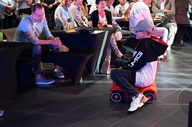 Ein Junge im schwarzen AMG-Hoodie und Mercedes-Cap fährt lachend auf einem roten Bobbycar durch den Showroom. Erwachsene Gäste sitzen an Tischen und beobachten ihn amüsiert. (Foto: Buchardt | Autohaus Peter )