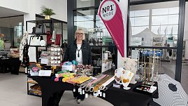 Stand von No1 Mode im Autohaus Autowelt Peter. Eine Frau präsentiert Taschen, Tücher, Schmuck und Accessoires, die auf Tischen und Regalen dekoriert sind. Daneben steht eine pinkfarbene Werbefahne mit dem Logo des Modegeschäfts. (Foto: Autohaus Peter)
