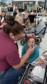Kinderschminken im Autohaus Autowelt Peter: Ein Mädchen mit Katzenohren-Stirnband bekommt ein buntes Regenbogen-Gesicht gemalt. Im Hintergrund sitzen weitere Familien mit Kindern. (Foto: Autohaus Peter)