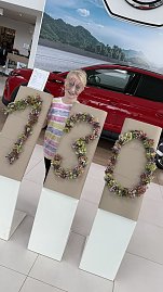„Ein Kind mit bunt bemaltem Gesicht steht im Autohaus Autowelt Peter hinter großen Ziffern aus Blumen, die 130 formen  passend zum Jubiläum 130 Jahre koda. Daneben ist ein rotes Auto ausgestellt. (Foto: Autohaus Peter)