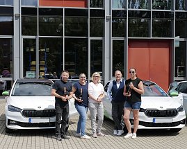 Fünf Personen stehen vor einem modernen Gebäude und posieren mit Champagnerflaschen vor zwei weißen koda-Fahrzeugen. Auf den Autos sind grüne Elemente und das Logo der „Praxisklinik für Zahnheilkunde“ zu sehen. (Foto: Autohaus Peter)