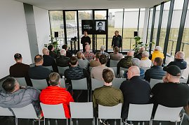 Eine Gruppe von Menschen sitzt in einem modernen, hellen Raum während einer Präsentation. Im Vordergrund sieht man mehrere Männer, die auf Stühlen sitzen und auf eine Bühne blicken. Auf der Bühne stehen zwei Personen: Ein Mann trägt einen schwarzen Pullover und spricht, während der andere in einem schwarzen Outfit steht. Auf einem Bildschirm im Hintergrund wird ein Porträt von Moritz Bleibtreu gezeigt. Der Raum hat große Fenster, durch die man die Natur draußen sieht. Pflanzen stehen in den Ecken des Raumes. (Foto: © 2025 HANSE VISUALS)