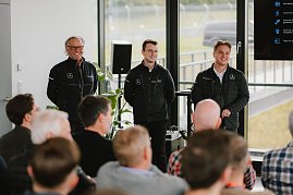 Drei Männer in schwarzen Jacken mit dem Mercedes-Benz 'Driving Experience'-Logo stehen vor einer Personengruppe und sprechen zu einem Publikum. Der Raum hat große Fenster, durch die man die Rennstrecke sieht. (Foto: © 2025 HANSE VISUALS)