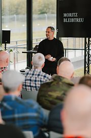 Ein Mann in schwarzem Pullover hält eine Tasse in der Hand und spricht zu einem Publikum. Auf dem Bildschirm hinter ihm ist der Name 'Moritz Bleibtreu' zu sehen. Die Zuschauer im Vordergrund sind nur von hinten zu sehen. (Foto: © 2025 HANSE VISUALS)