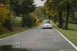 Eine Gruppe von Fahrzeugen fährt auf einer kurvigen Landstraße, die von Bäumen gesäumt ist. Im Vordergrund ist ein weißer Mercedes-Benz Cabrio zu sehen, gefolgt von weiteren Fahrzeugen. Die Bäume zeigen herbstliche Farben, und die Straße ist von grünen Wiesen und einem hügeligen Hintergrund umgeben. (Foto: © 2025 HANSE VISUALS)