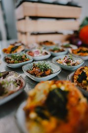 Mehrere kleine Schalen mit verschiedenen Gerichten sind auf einem Tisch arrangiert. Die Schalen enthalten bunte, frisch zubereitete Speisen, die mit Kräutern garniert sind. Im Hintergrund ist ein Stapel von Holzkisten sichtbar und einige weitere Speisen sind unscharf zu sehen. (Foto: © 2025 HANSE VISUALS)