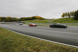 Mehrere Autos fahren auf einer Rennstrecke. Im Vordergrund ist ein schwarzer Mercedes-AMG und ein oranger Mercedes-AMG GT zu sehen. Weitere Fahrzeuge sind in der Ferne sichtbar, während die Strecke von Bäumen gesäumt ist, die herbstliche Farben zeigen. Der Himmel ist bewölkt, und die Strecke führt in eine Kurve. (Foto: © 2025 HANSE VISUALS)
