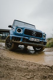 Ein blauer Mercedes-Benz G-Class fährt auf einem matschigen Gelände. Der Blickwinkel zeigt das Fahrzeug aus der Nähe, mit dem Fokus auf die Frontpartie und die markante Kühlergrill. Der Unterboden und die Räder sind mit Schlamm bedeckt, was die Geländetauglichkeit des Fahrzeugs betont. Im Hintergrund ist ein Gebäude unscharf zu sehen. (Foto: © 2025 HANSE VISUALS)