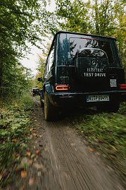 Ein Mercedes-Benz G-Class, Modell G 580, fährt auf einem unbefestigten Waldweg. Das Fahrzeug ist mit dem Aufkleber 'ELECTRIC TEST DRIVE' auf der Heckklappe versehen. Die Reifen sind mit Schlamm bedeckt, und im Hintergrund ist ein weiteres Fahrzeug zu sehen. Der Wald mit grünen Bäumen umgibt den Weg. (Foto: © 2025 HANSE VISUALS)