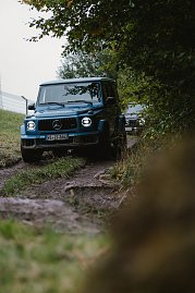 Ein blauer Mercedes-Benz G-Class fährt auf einem unbefestigten, felsigen Weg im Wald. Der Blick ist von der Seite, sodass die Vorderseite des Fahrzeugs und die charakteristischen Scheinwerfer gut sichtbar sind. Im Hintergrund ist ein weiteres Fahrzeug zu sehen. Der Weg ist von Gras und Bäumen gesäumt. (Foto: © 2025 HANSE VISUALS)