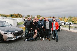 Eine Gruppe von Männern steht lachend zwischen zwei Mercedes-Maybach Fahrzeugen auf dem Gelände der Mercedes-Benz Driving Experience am Bilster Berg. Einige tragen bunte Jacken, und einer sitzt entspannt auf dem Boden. Im Hintergrund sind Gebäude der Rennstrecke und herbstlich gefärbte Bäume zu sehen. (Foto: © 2025 HANSE VISUALS)
