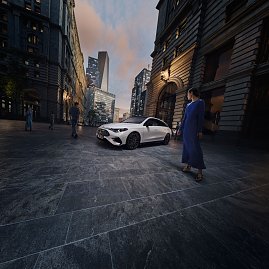 Das Bild zeigt einen wei&szlig;en Mercedes-Benz CLA Shooting Brake, der auf einer breiten, nahezu leeren Stadtstra&szlig;e f&auml;hrt. Moderne Geb&auml;ude umgeben die Szene, das Licht wirkt weich und fr&uuml;hmorgendlich. Der Wagen ist seitlich zu sehen und vermittelt Dynamik sowie Eleganz.