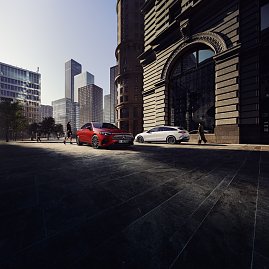 Das Bild zeigt einen wei&szlig;en Mercedes-Benz CLA Shooting Brake, der auf einer breiten, nahezu leeren Stadtstra&szlig;e f&auml;hrt. Moderne Geb&auml;ude umgeben die Szene, das Licht wirkt weich und fr&uuml;hmorgendlich. Der Wagen ist seitlich zu sehen und vermittelt Dynamik sowie Eleganz.