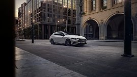 Das Bild zeigt einen wei&szlig;en Mercedes-Benz CLA Shooting Brake, der auf einer breiten, nahezu leeren Stadtstra&szlig;e f&auml;hrt. Moderne Geb&auml;ude umgeben die Szene, das Licht wirkt weich und fr&uuml;hmorgendlich. Der Wagen ist seitlich zu sehen und vermittelt Dynamik sowie Eleganz.