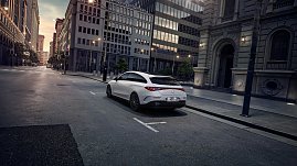 Das Bild zeigt einen wei&szlig;en Mercedes-Benz CLA Shooting Brake, der auf einer breiten, nahezu leeren Stadtstra&szlig;e f&auml;hrt. Moderne Geb&auml;ude umgeben die Szene, das Licht wirkt weich und fr&uuml;hmorgendlich. Der Wagen ist seitlich zu sehen und vermittelt Dynamik sowie Eleganz.
