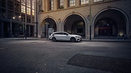 Das Bild zeigt einen wei&szlig;en Mercedes-Benz CLA Shooting Brake, der auf einer breiten, nahezu leeren Stadtstra&szlig;e f&auml;hrt. Moderne Geb&auml;ude umgeben die Szene, das Licht wirkt weich und fr&uuml;hmorgendlich. Der Wagen ist seitlich zu sehen und vermittelt Dynamik sowie Eleganz.