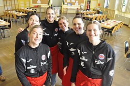 6 junge Mädchen mit schwarzen Sportoberteilen und roten Schürzen lächeln in die Kamera.