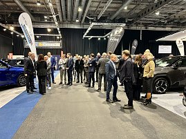 Viele Messebesucher stehen zwischen ausgestellten Autos und Markenflaggen und unterhalten sich in der Ausstellungshalle.