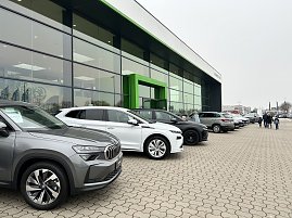 Reihe geparkter Autos vor einem modernen Autohaus mit gro&szlig;er Glasfront und gr&uuml;nem Eingang, Menschen gehen &uuml;ber den Parkplatz.